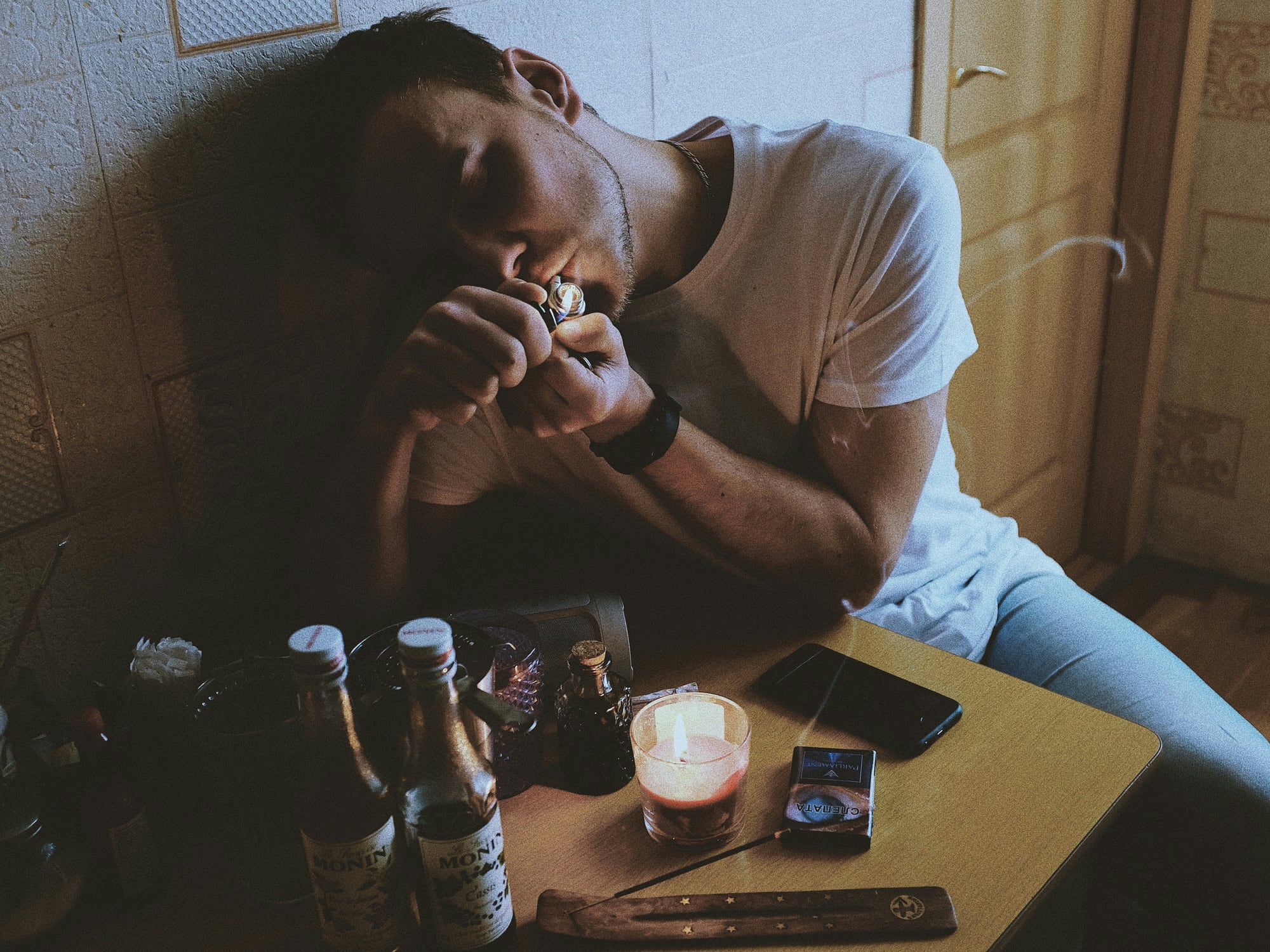 A man slumped at a table beside bottles, a lit candle and cigarettes