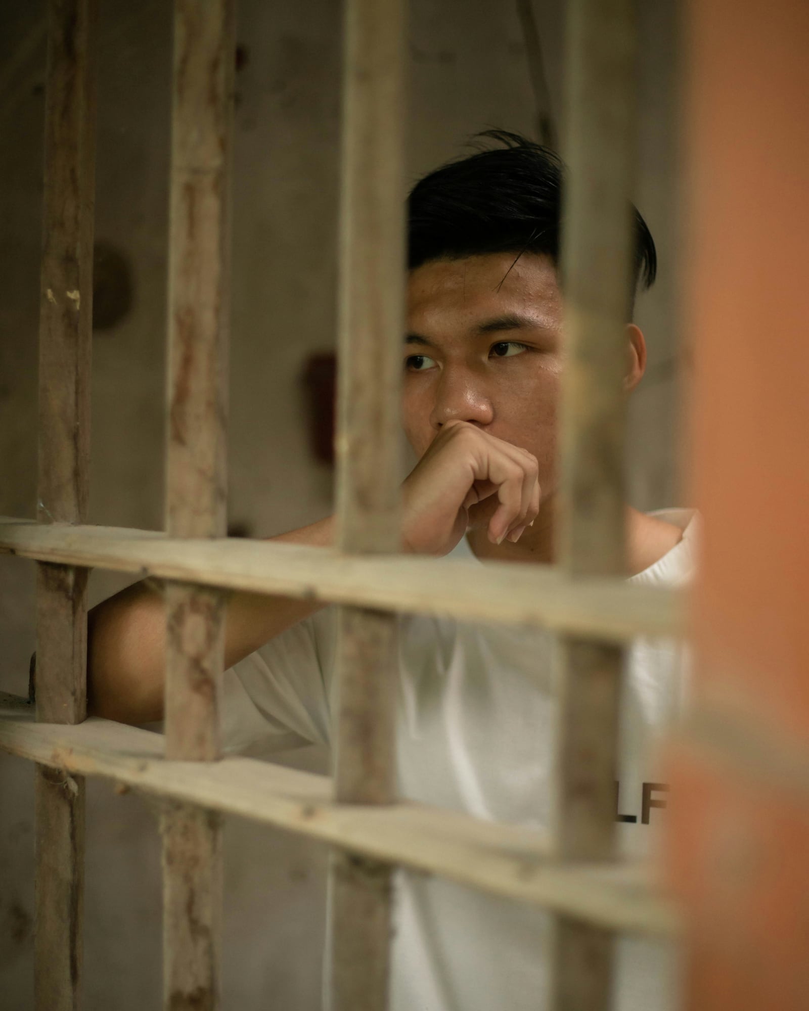 A young man behind wooden bars, hand raised to his mouth, looking to the side